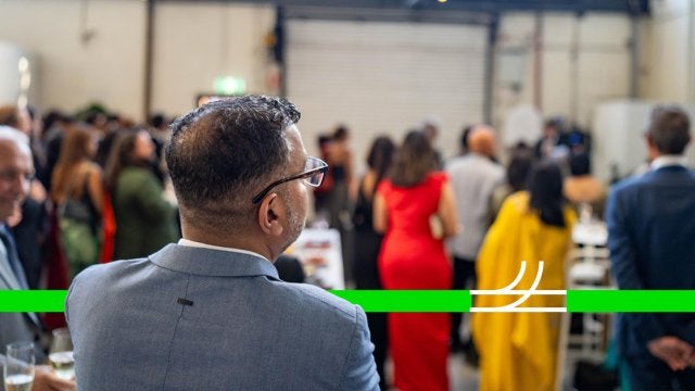 Ejecutivo observando un evento corporativo desde la audiencia en un entorno profesional y elegante.